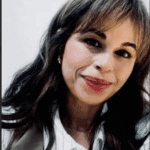 A social worker with long brown hair, wearing a white blouse, smiles at the camera against a plain background.