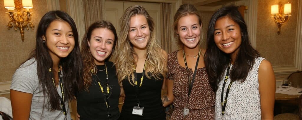 Five young women smiling and standing together indoors, all wearing name badges and conference lanyards.
