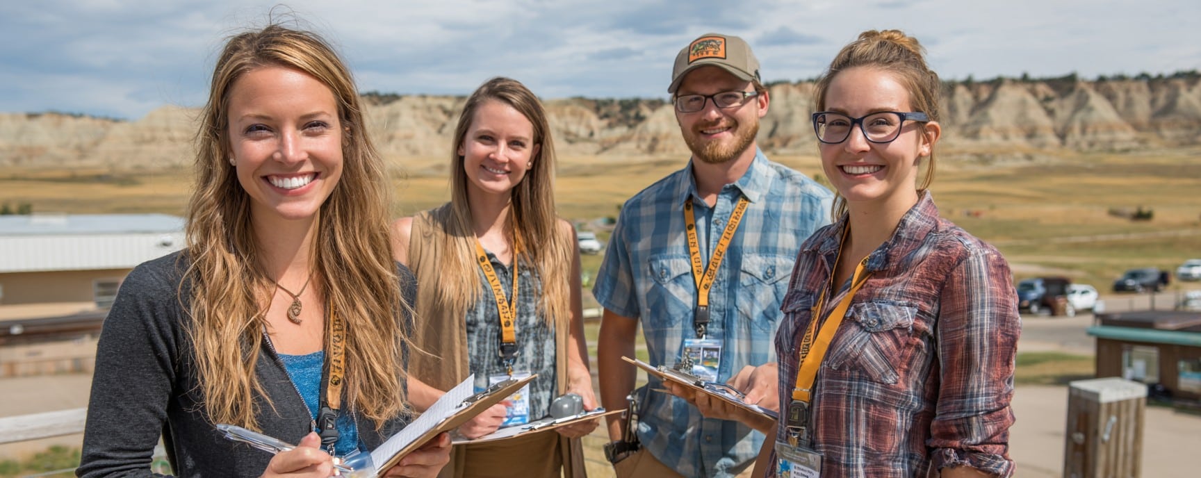 Four smiling young adults with clipboards stand outdoors in a rural area, wearing name tags and casual clothes.