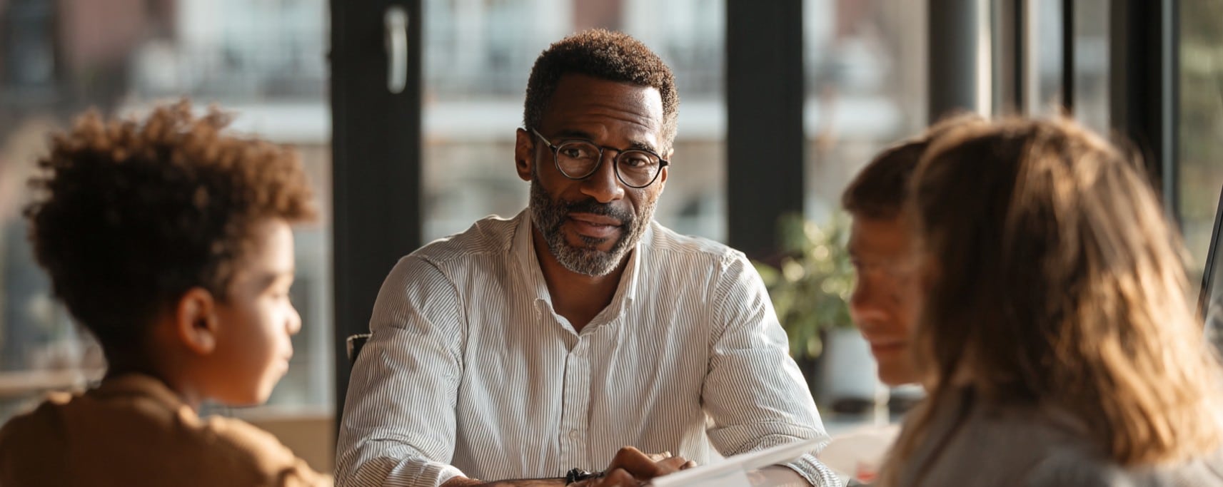 A man in glasses sits at a table with three children, having a serious conversation in a sunlit room.