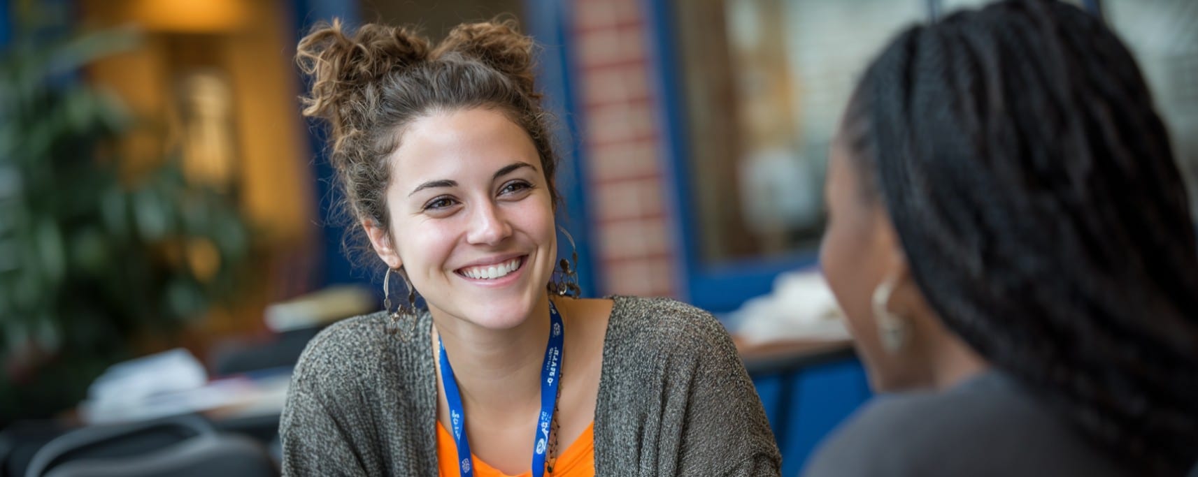 A young woman with curly hair smiles while talking to another person indoors.