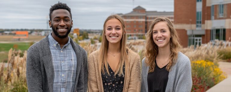 Three young adults stand outside, smiling, with buildings and greenery in the background.