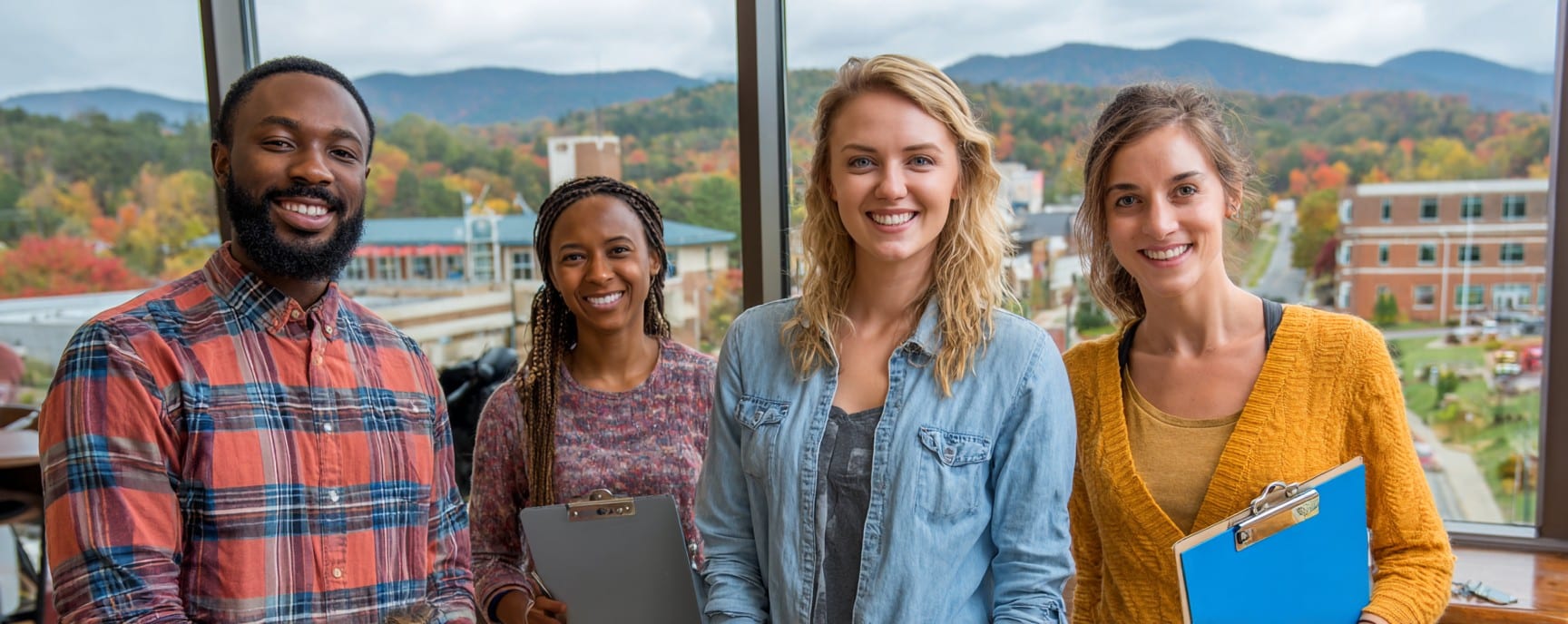 Four smiling young adults holding clipboards stand indoors with a campus and mountains visible through the window.