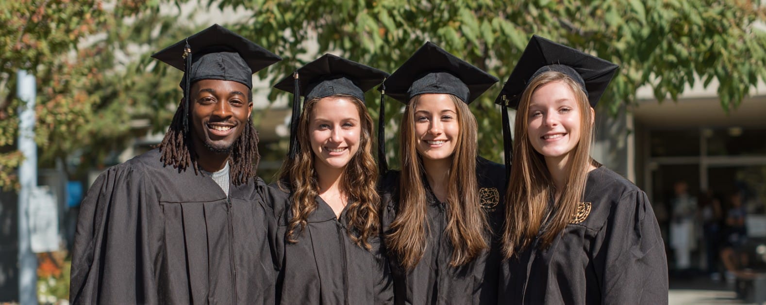 Four social work graduates in caps and gowns smile together outdoors in front of a leafy building.